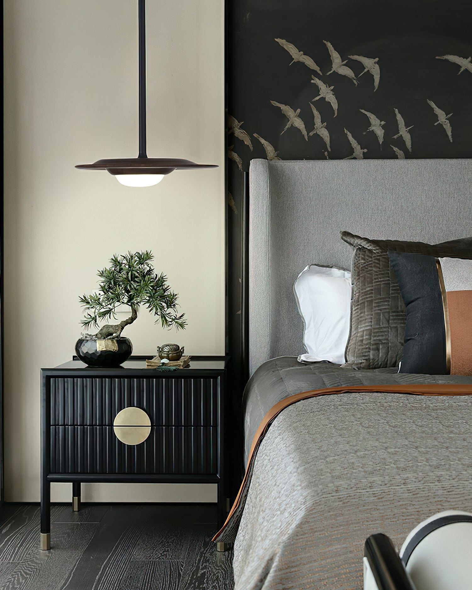 Dark wood-tone pendant beside a bed, above a black ribbed cabinet with a round gold handle and bonsai