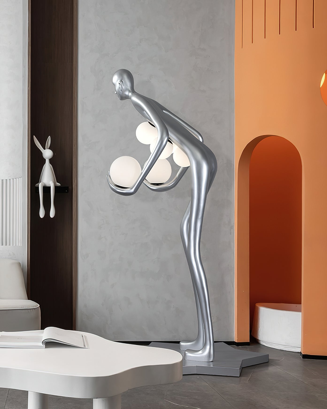 Silver figure seen from behind beside orange arch niches, a white table and rabbit ornament