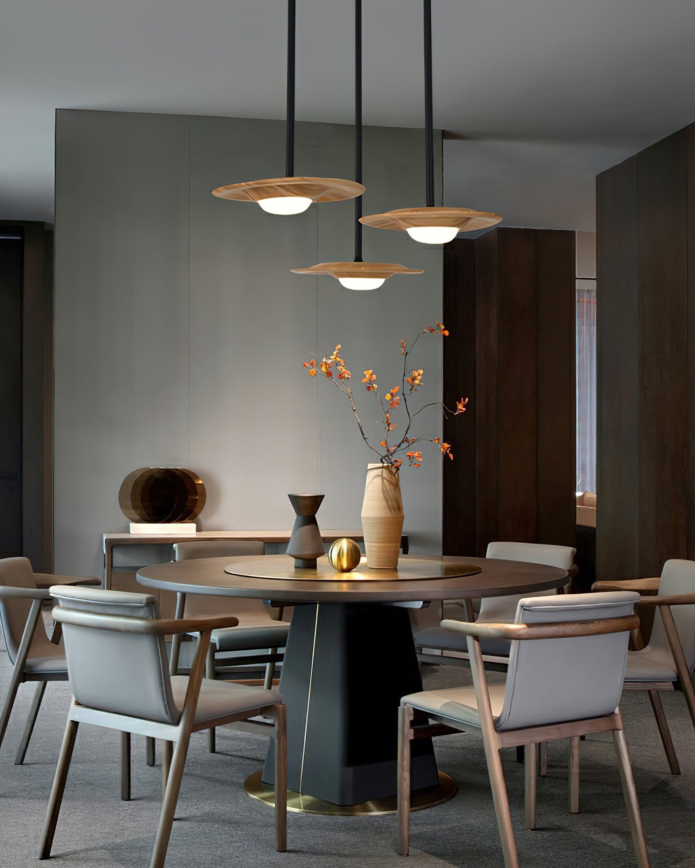 Three light wood-grain pendants at staggered heights above a round dining table with a tall vase and upholstered chairs