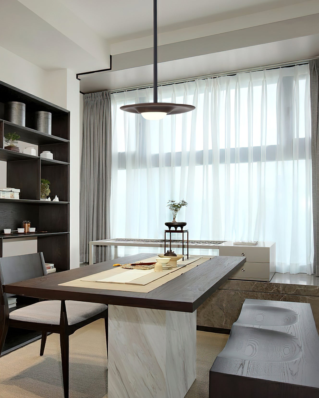 Dark wood-tone disc pendant with glowing white dome above a wood table beside sheer curtains and dark open shelving
