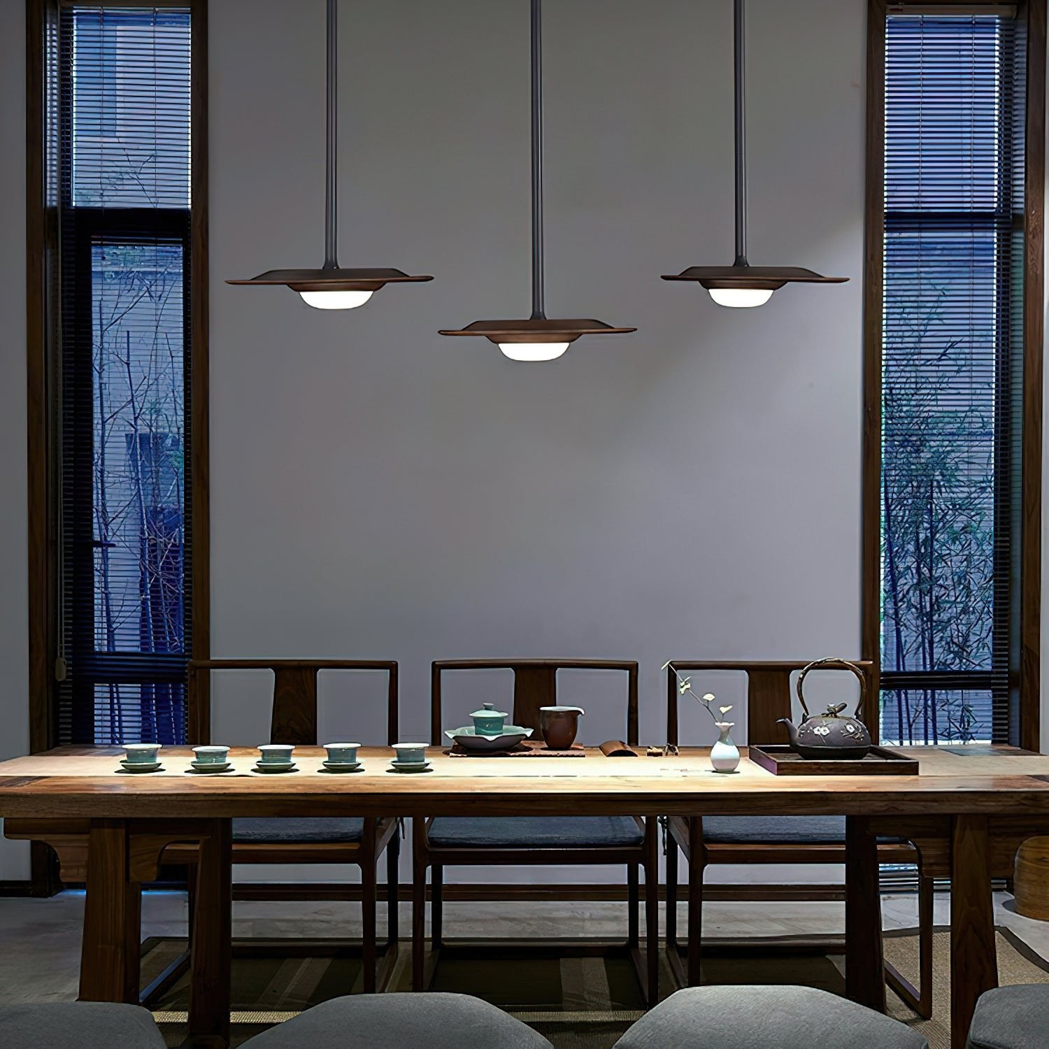 Three dark wood-tone disc pendant shades above a long tea table with cups, teapots and wood chairs between window blinds