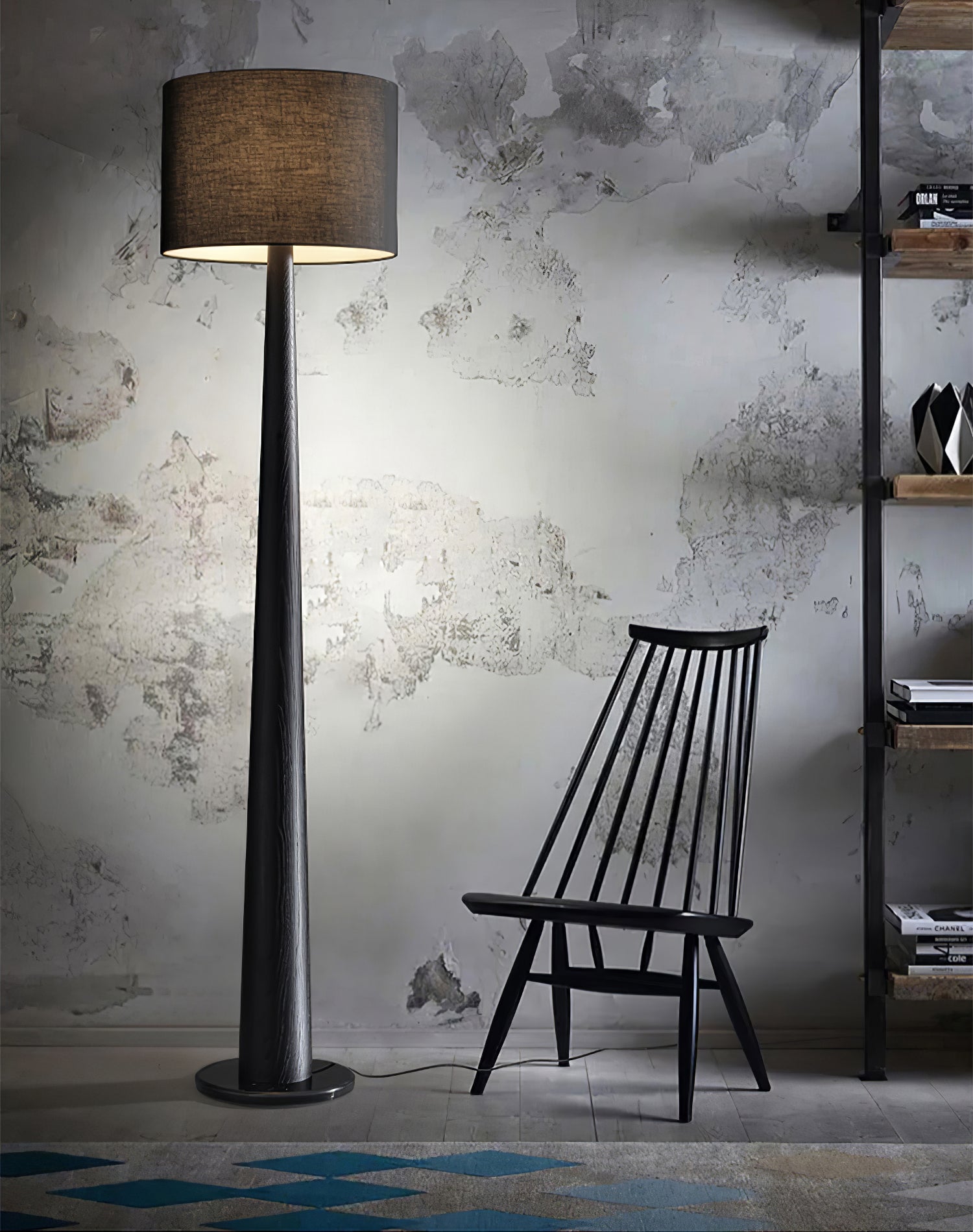 Black tapered floor lamp with dark drum shade beside a black spindle-back chair and shelves against a textured wall