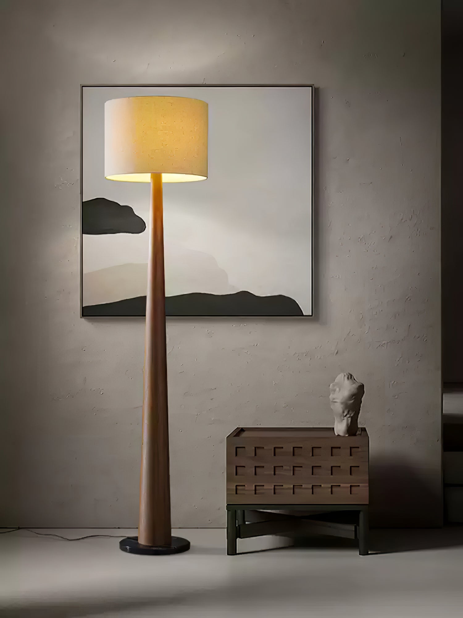 Wood-tone tapered floor lamp with illuminated drum shade beside a low cabinet and abstract framed artwork