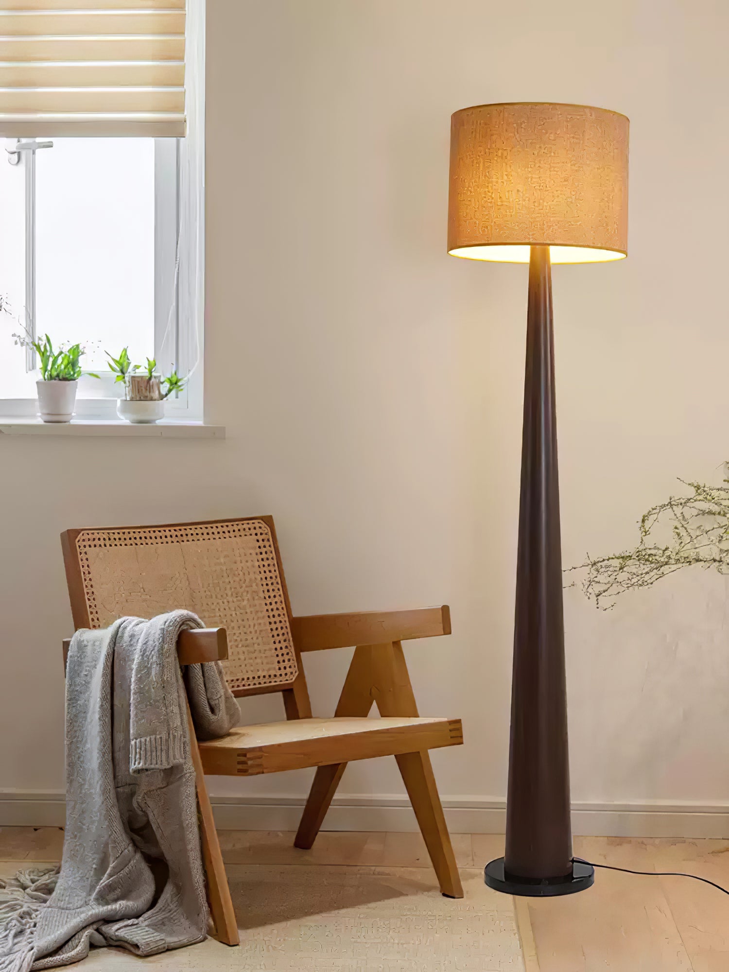 Dark brown floor lamp with textured beige shade beside a cane-back armchair, gray throw and small window