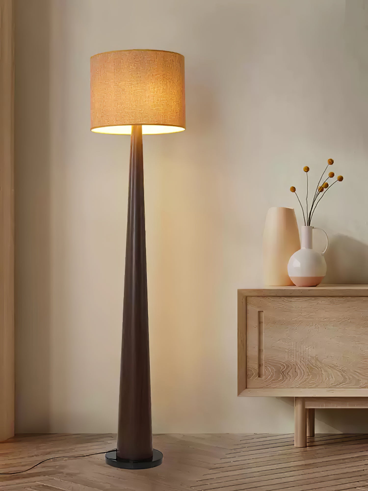 Dark brown tapered floor lamp with textured beige shade beside a light carved cabinet and white vases