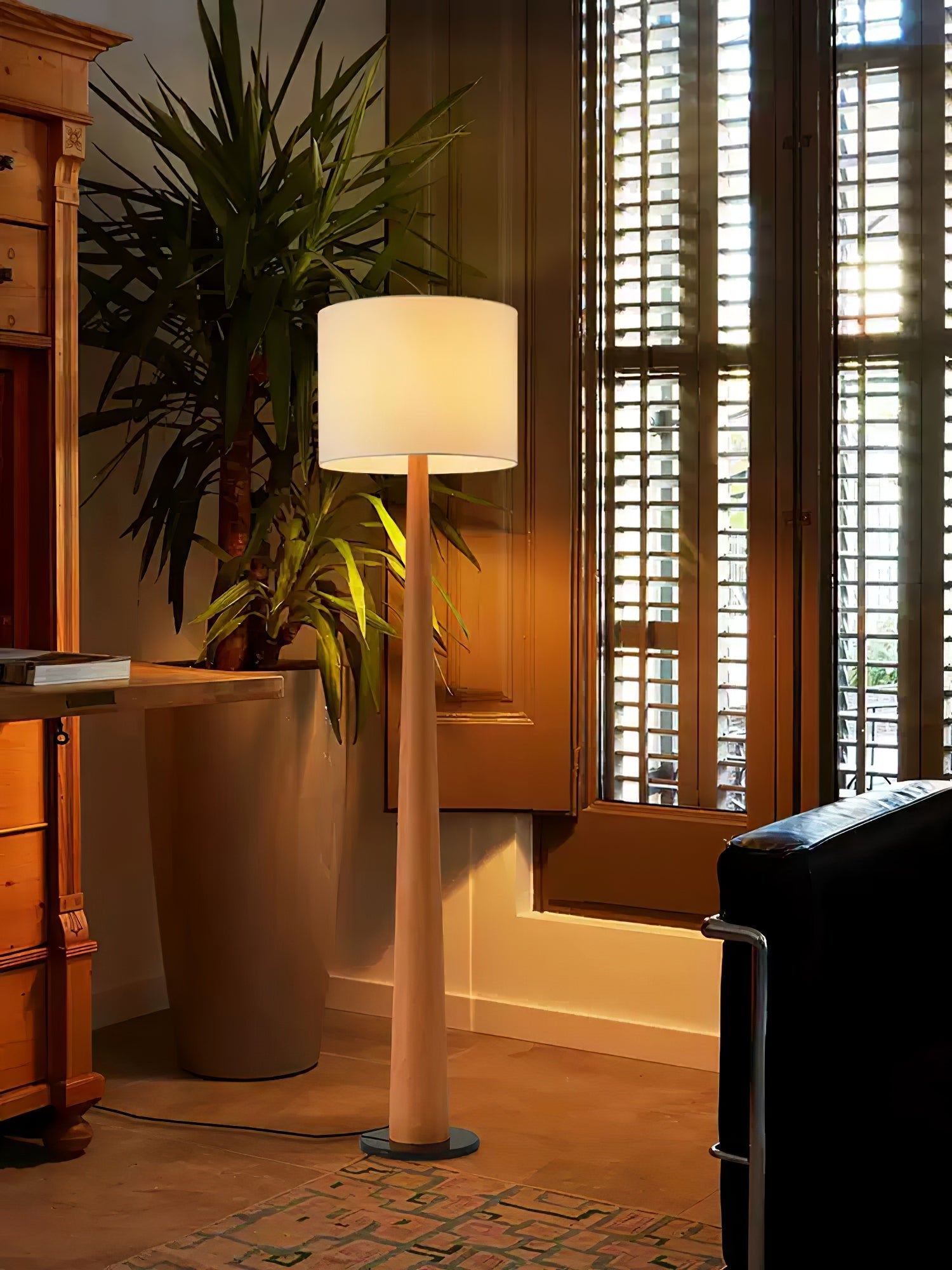 Light wood-tone floor lamp with a pale drum shade beside a tall plant, wooden shutters and dark seating