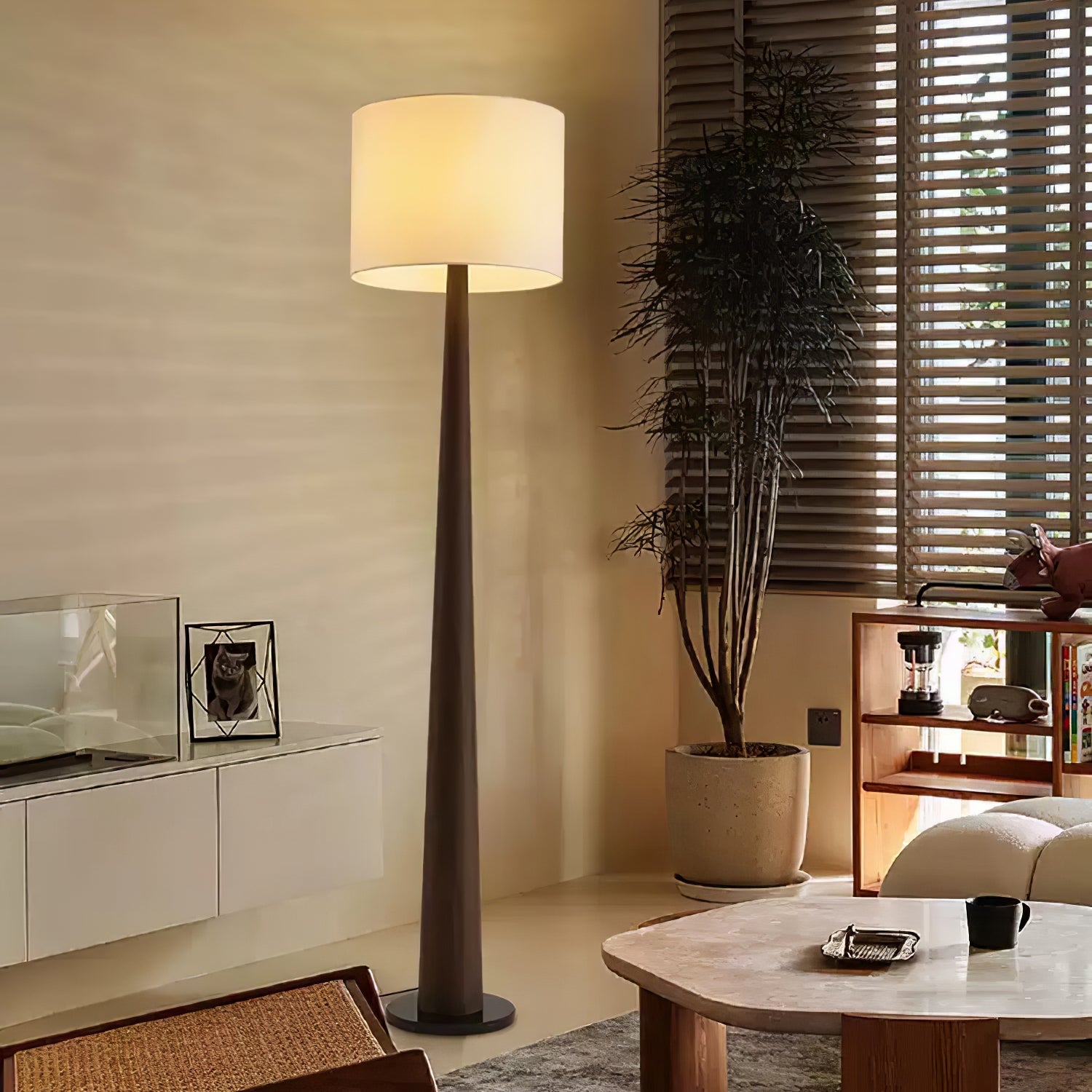 Brown tapered floor lamp with a pale illuminated shade beside a potted tree and wooden blinds