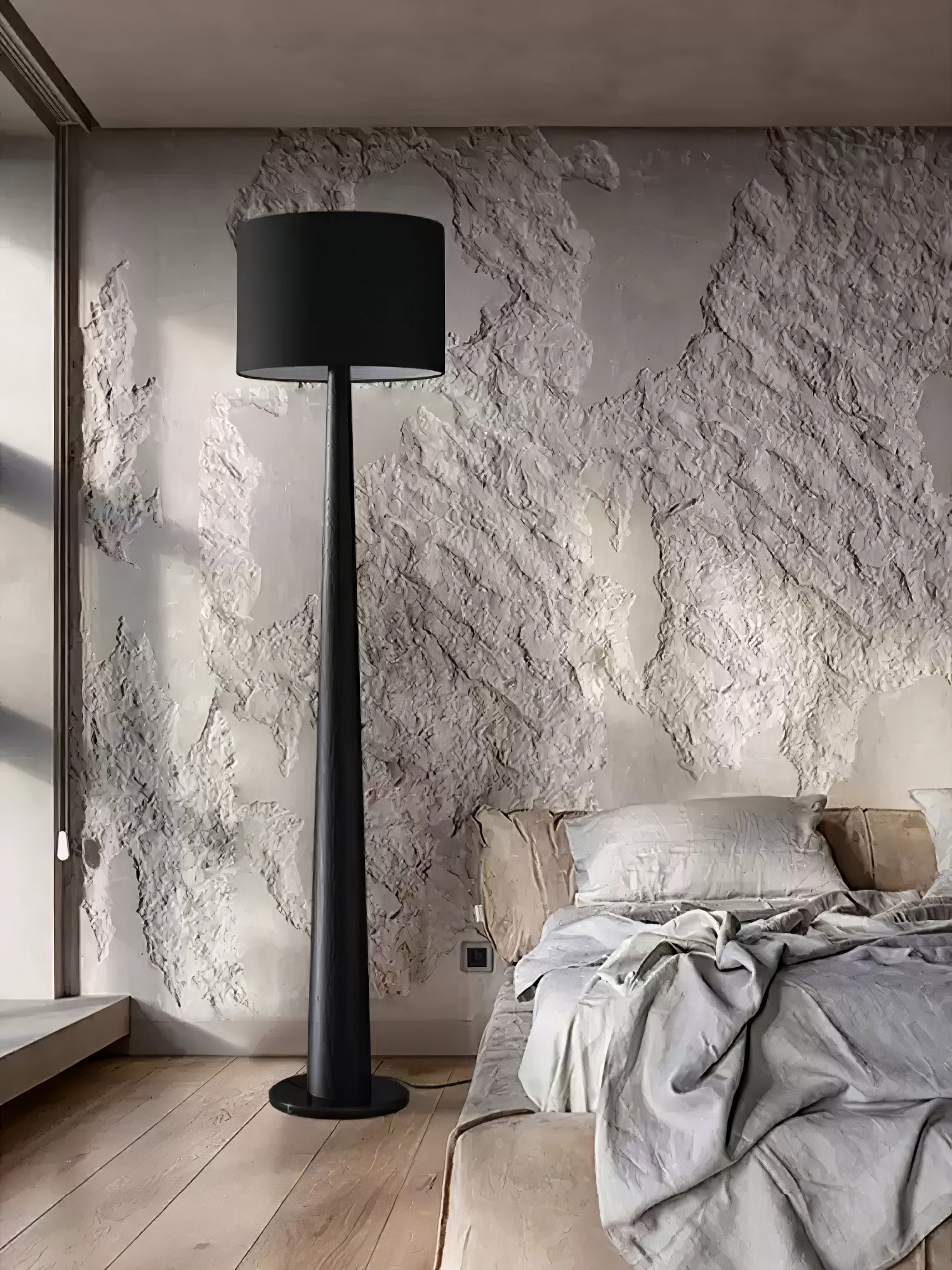 Black tapered floor lamp with black drum shade beside a bed against a textured pale wall