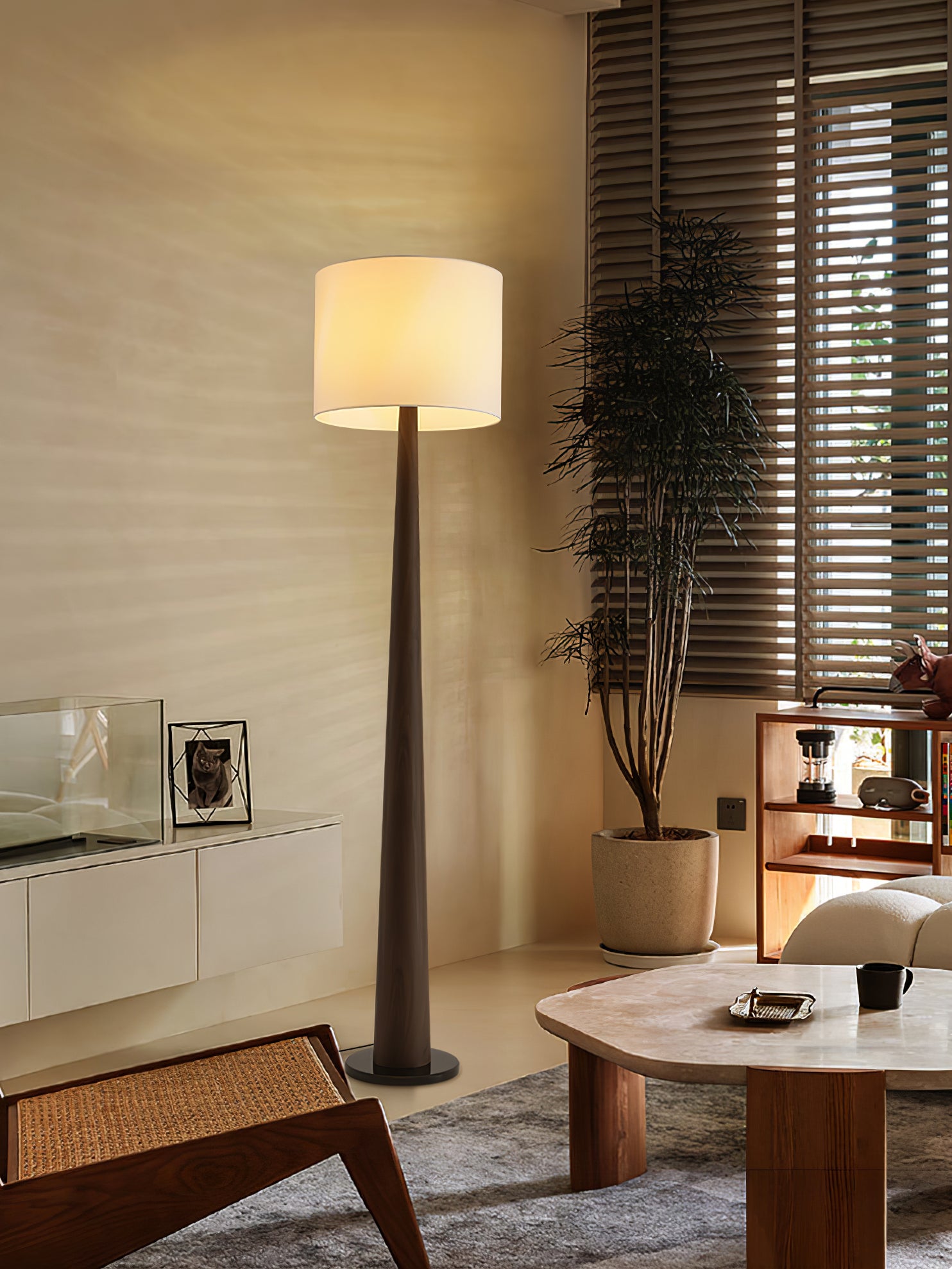 Brown floor lamp with pale illuminated shade beside a tree, blinds, white cabinet and coffee table