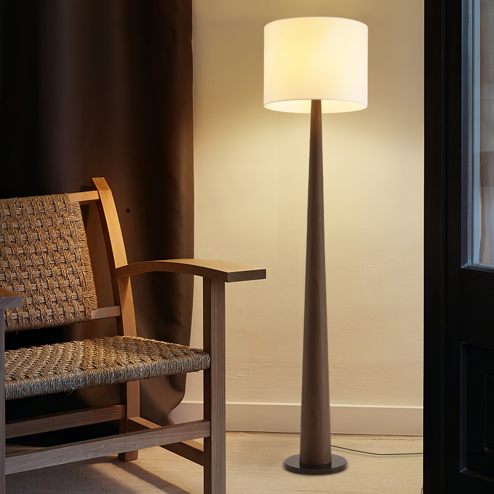 Brown tapered floor lamp with a pale drum shade beside a woven wooden armchair and window