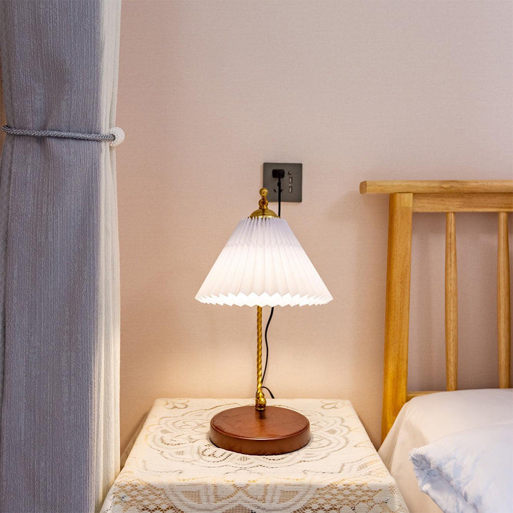 White-looking pleated table lamp on a lace-covered bedside table near a wood bedhead and grey curtains