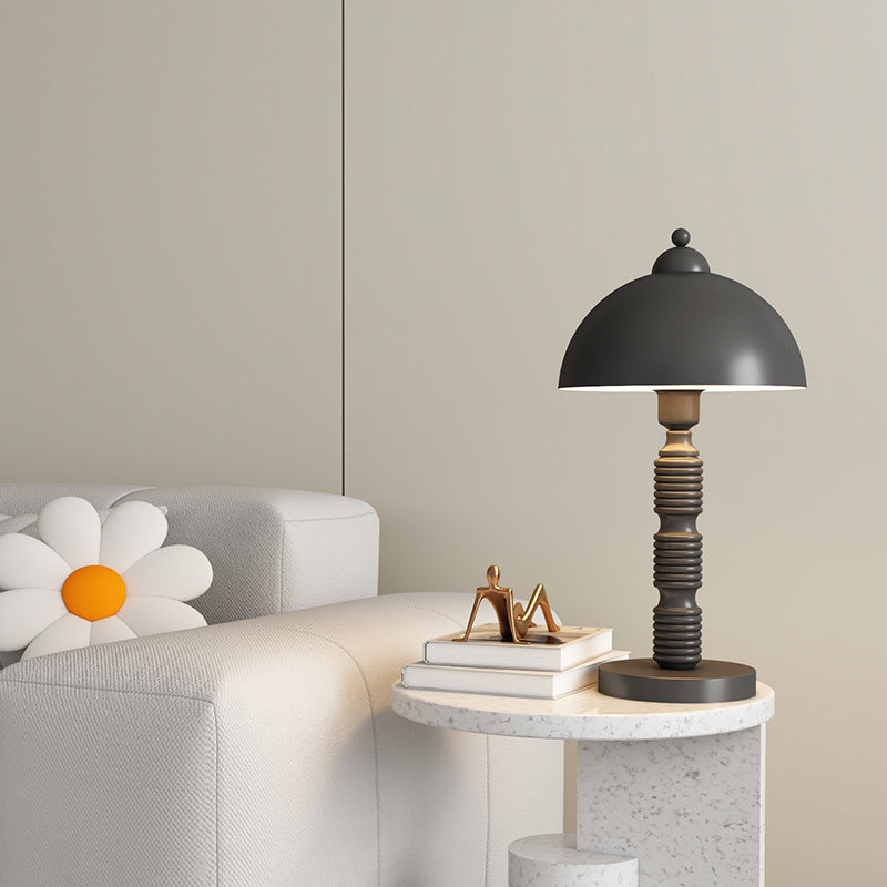 Black ribbed-stem lamp on a pale round side table beside books and a seated figurine, near a sofa with daisy cushion