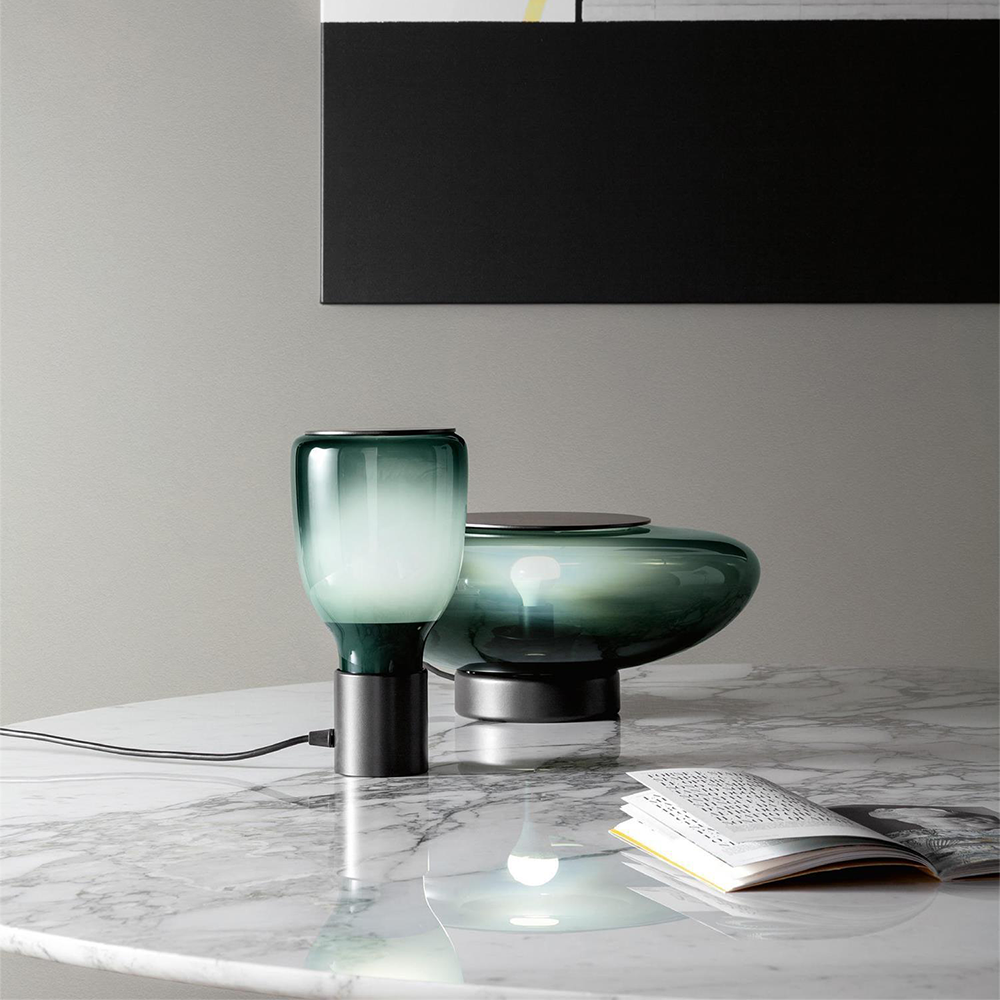 Tall green bottle-shaped lamp beside a wide green bowl-shaped lamp and an open book on a marble-look table