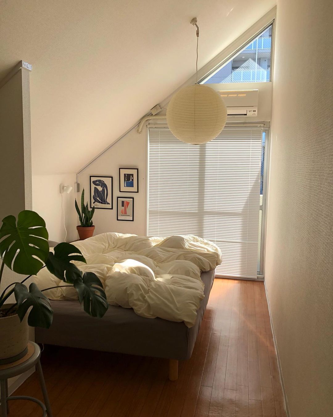 White globe above a bed under a pitched ceiling beside triangular window, framed art and plants