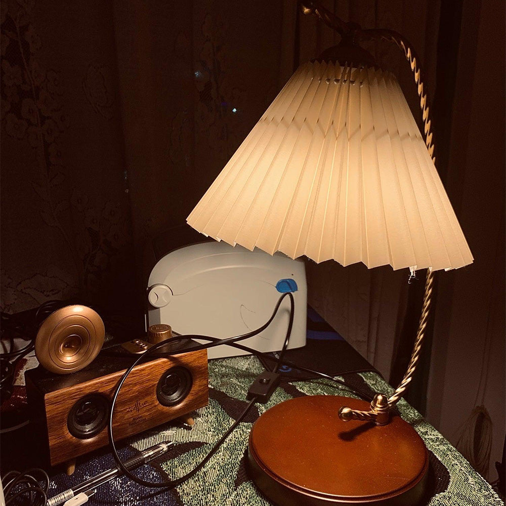 Lit pleated table lamp beside speakers on green patterned fabric, with a black cord and inline box