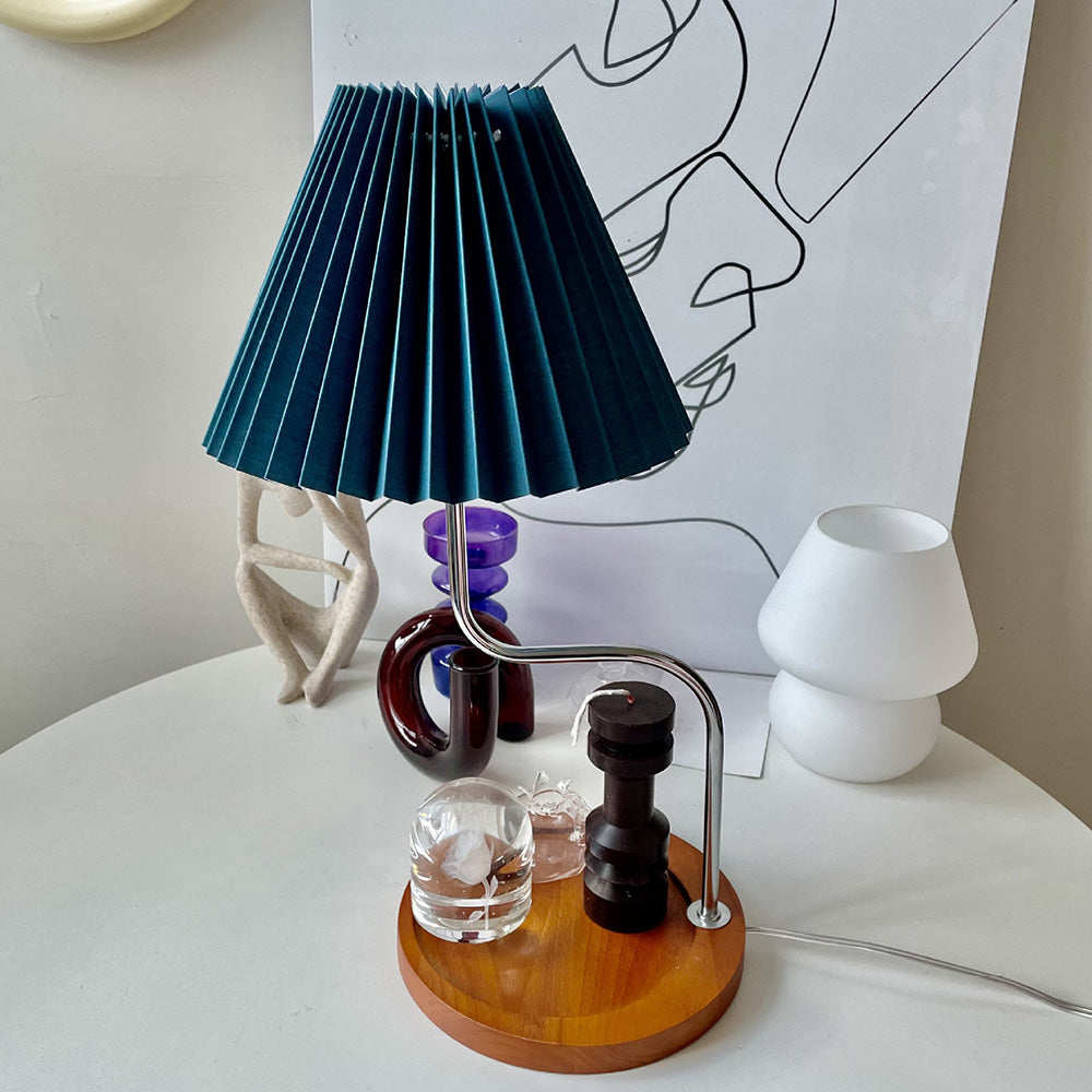 Blue pleated shade on bent silver-coloured stem and round wood-looking base holding decorative glass items and black candle