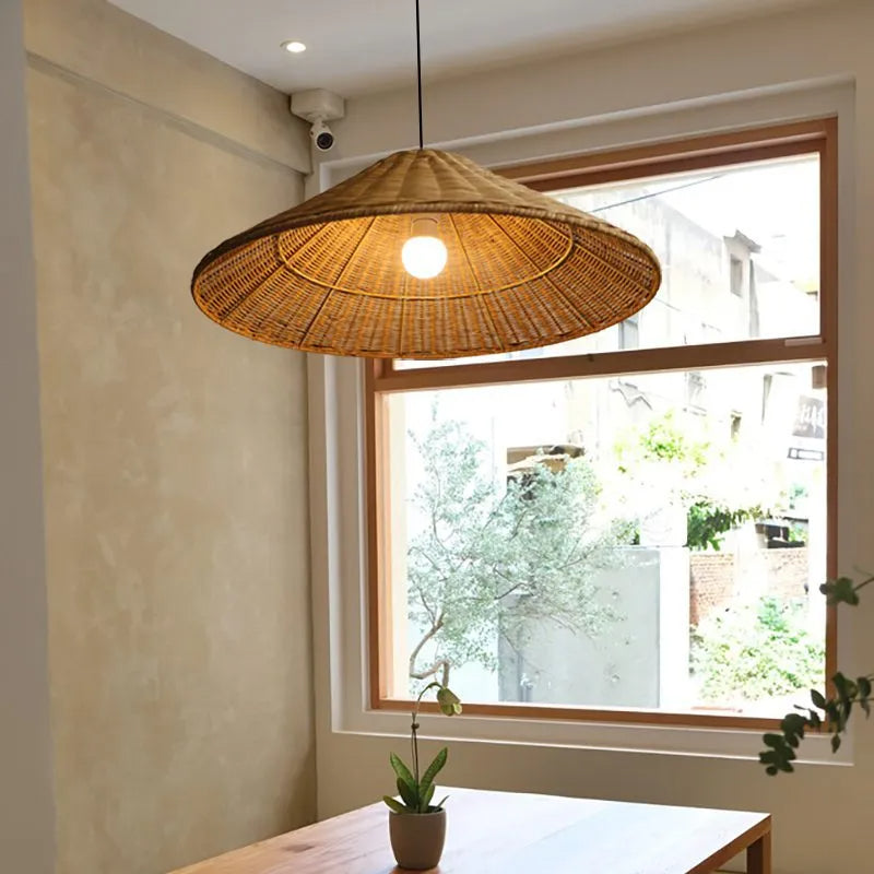 Brown woven cone shade with exposed lit bulb above a window-side wooden table and small potted plant