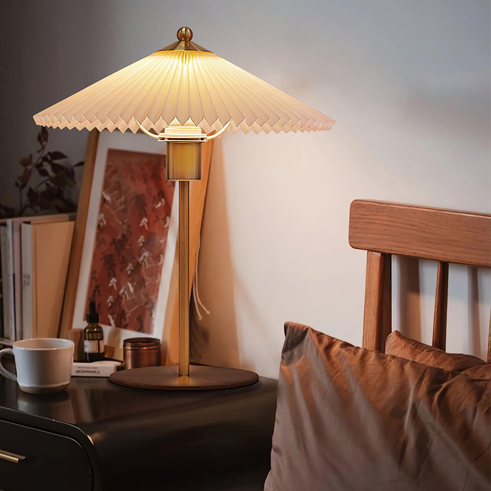 Lit pleated lamp beside a wooden headboard with brown bedding, mug and framed botanical art