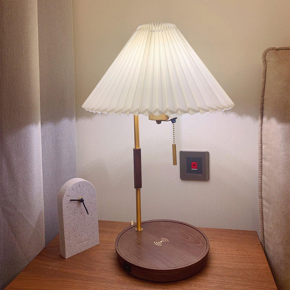 Lit pleated lamp with a charging symbol on its brown base beside a clock and beige headboard