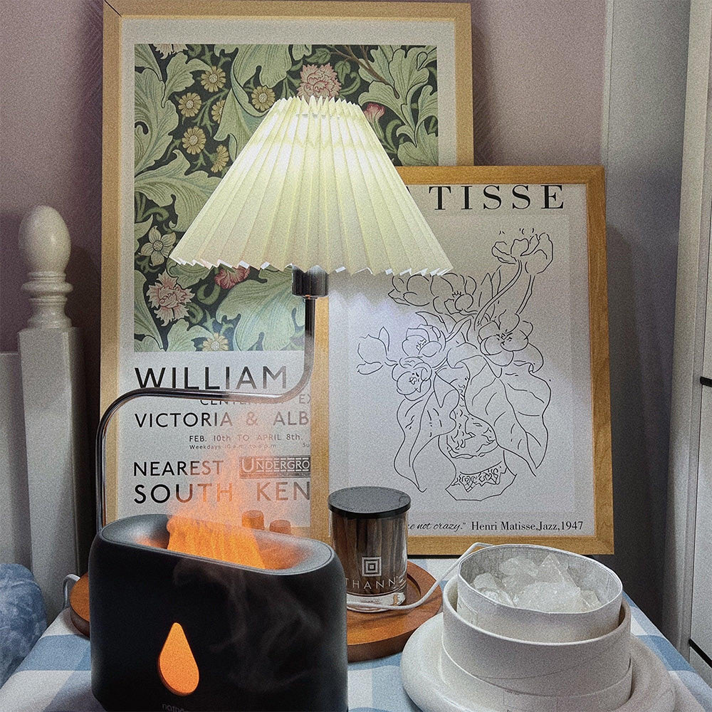 Lit pleated table lamp before floral and line-art prints, near a jar, bowls and a flame-looking appliance