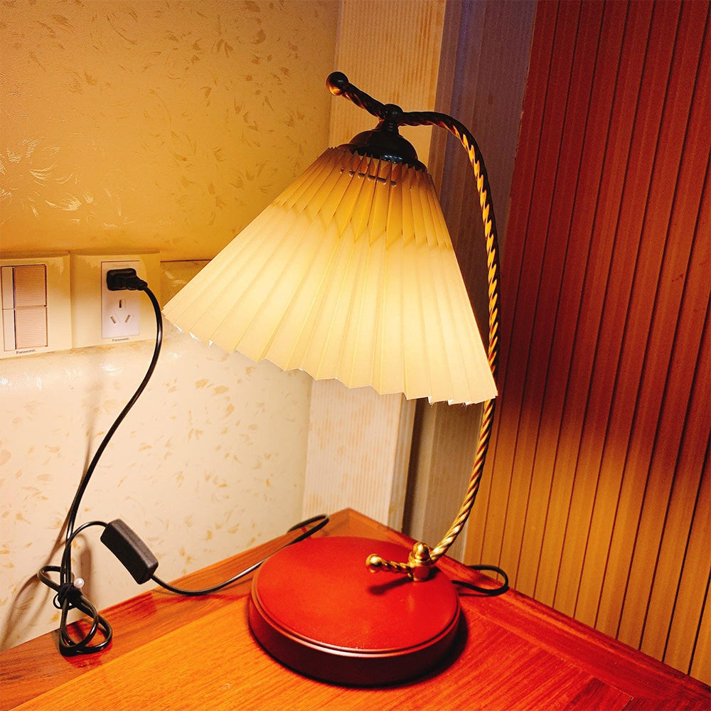 Pleated table lamp on a wooden bedside table, with black cord, inline box and plug at a wall socket
