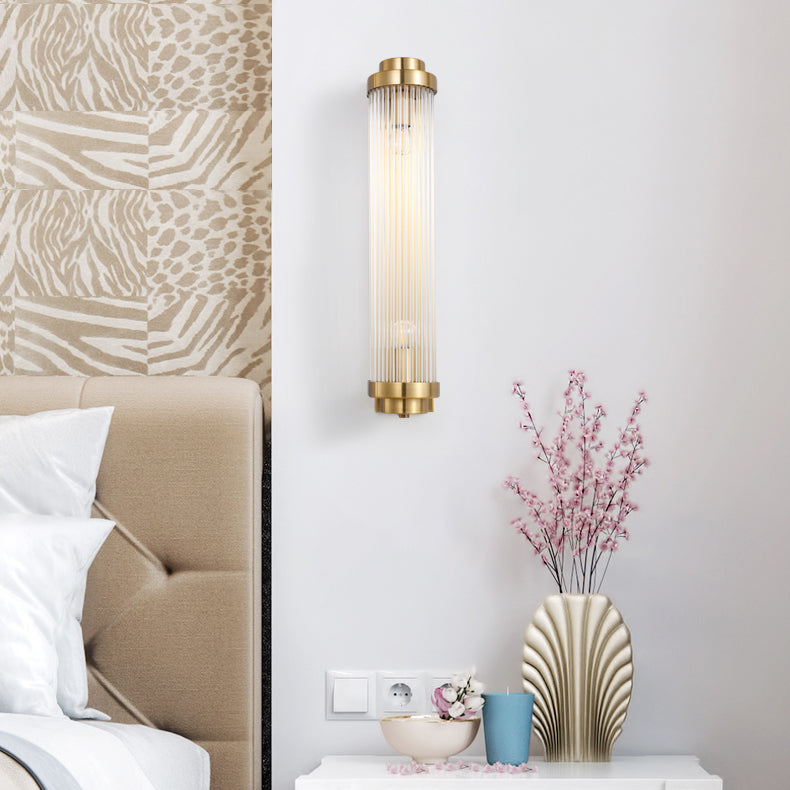 Gold rod wall light near a beige headboard, patterned wall, white pillows and a vase of pink stems beside a blue cup