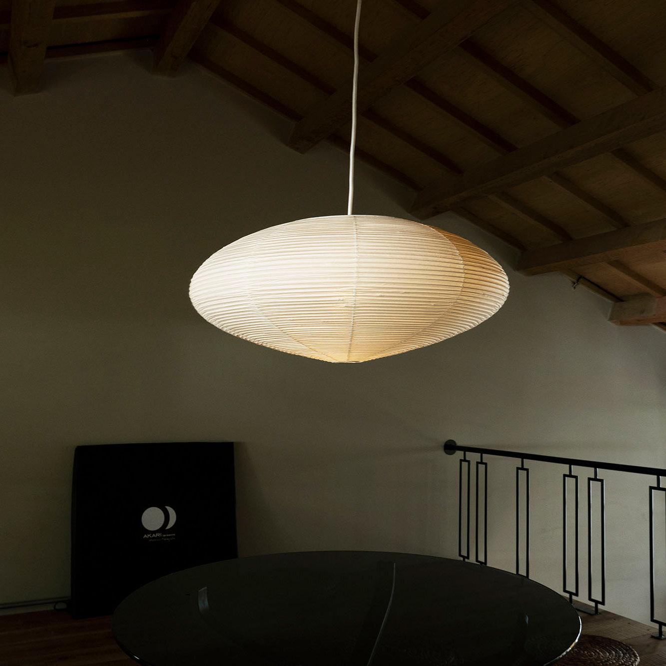 Glowing flattened paper shade above a round black table beside a railing under exposed timber beams