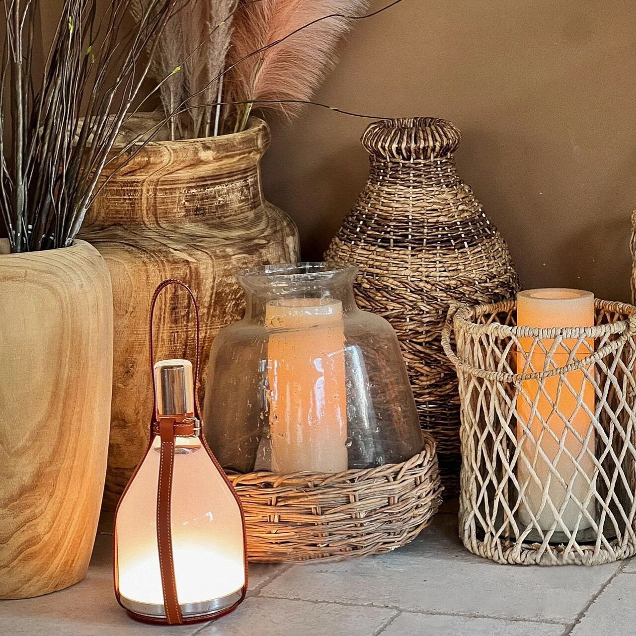 Small leather-handle Bell lamp among wood-grain vases, woven baskets, a glass candle cloche and dried pink plumes