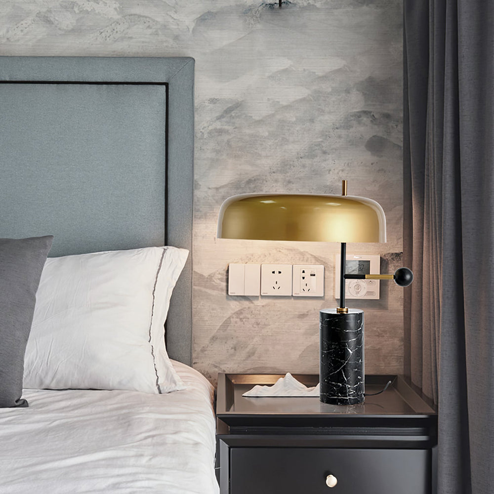Lit gold-shade lamp with black veined base on a bedside cabinet near white bedding and a dark curtain