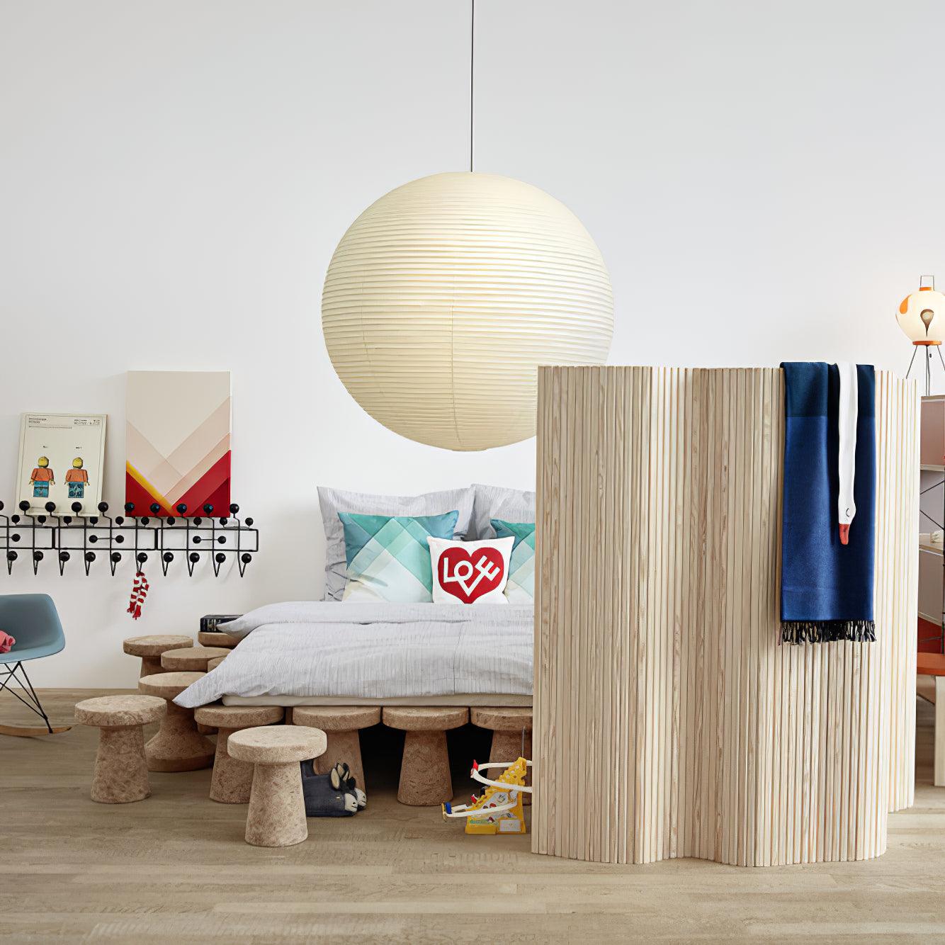 Paper globe above a grey-covered bed with heart pillow beside a wood screen, cork stools and rocking chair