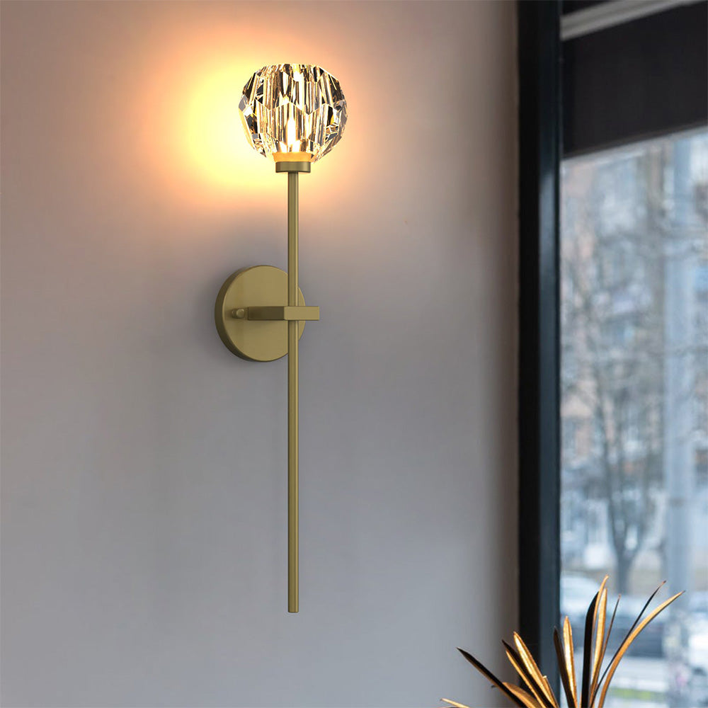 Lit faceted ball and long gold-tone stem on a pale wall by a dark-framed window and plant