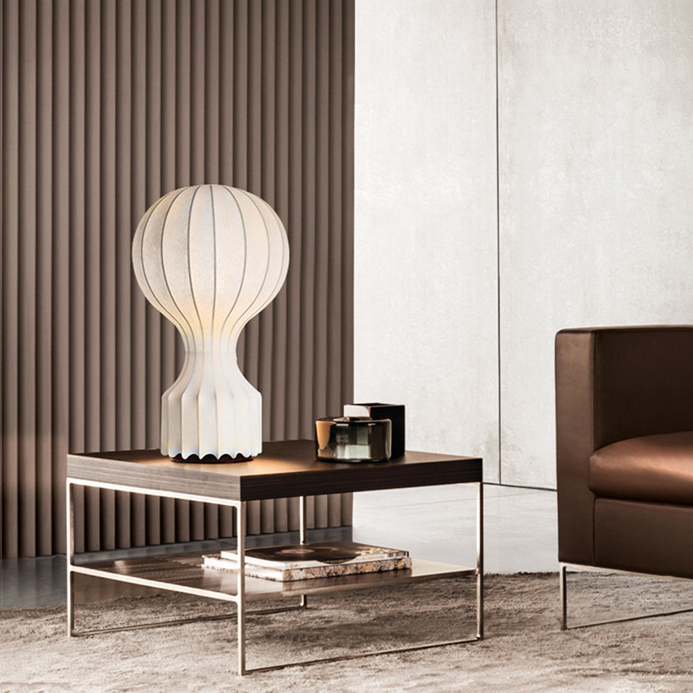 Lit Gatto lamp on a dark wooden low table with a silver-tone frame, a dark container and books on the shelf below
