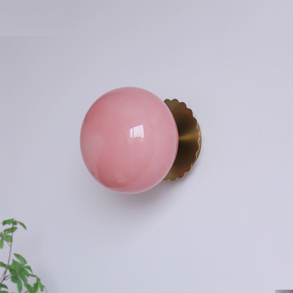 Pink glossy globe in front of gold-tone scalloped trim and round plate on a pale wall