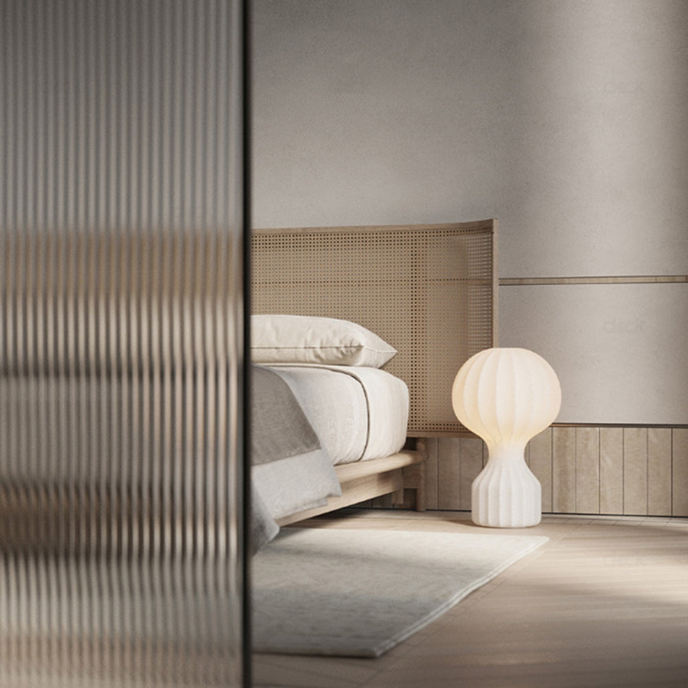 Illuminated ribbed lamp pictured beside a low bed with a woven headboard, pale bedding and rug, viewed past a textured partition