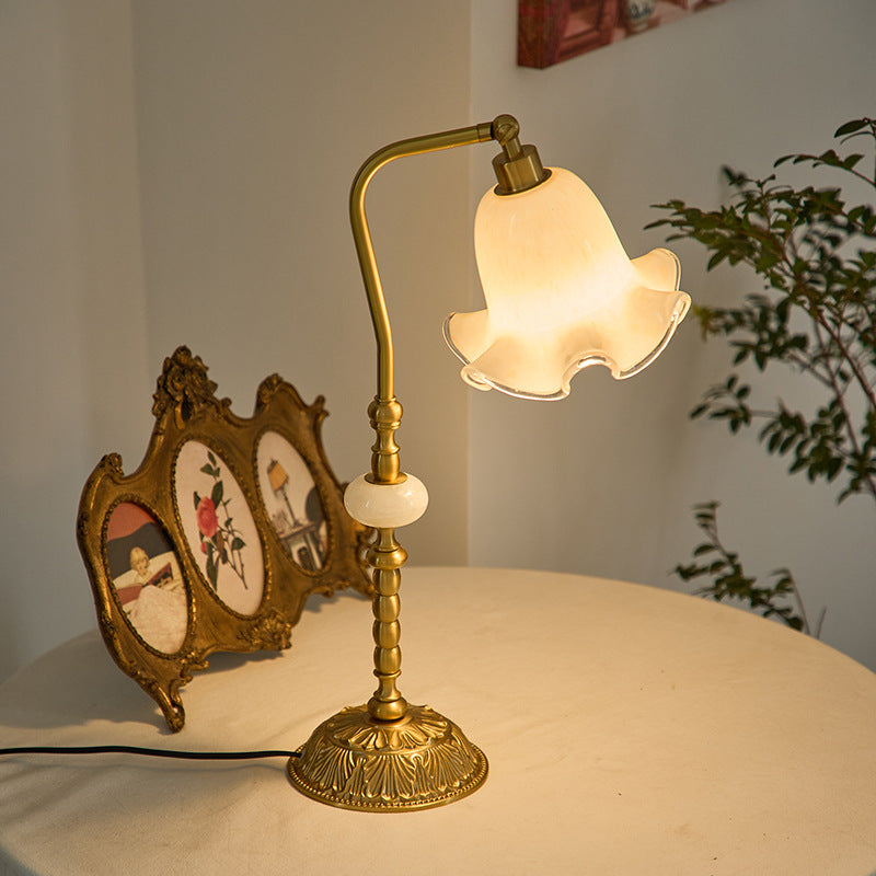 Illuminated cream flower-shade lamp beside an ornate triple frame and leafy plant on a pale cloth-covered table