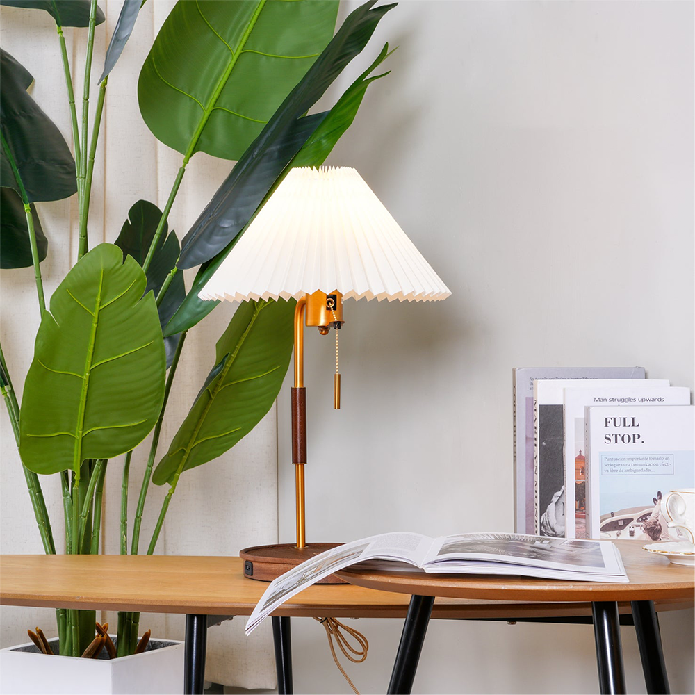 Glowing pleated table lamp on a wood desk beside a leafy plant, books and an open book