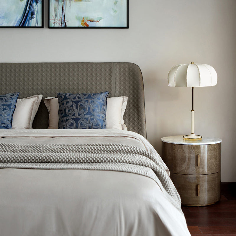 White-stem dome table lamp on round bedside cabinet beside gray quilted headboard and blue-and-white pillows