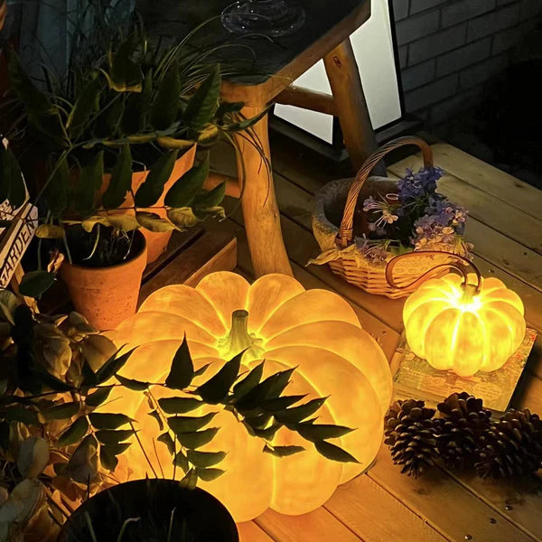 Large and small glowing pumpkin lamps on wood planks beside leafy plants, flower basket and pinecones