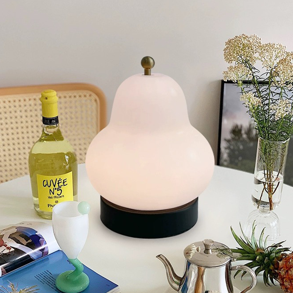 Glowing pear-shaped white lamp among a wine bottle, flower vase, books, green-based cup and silver teapot on a pale table