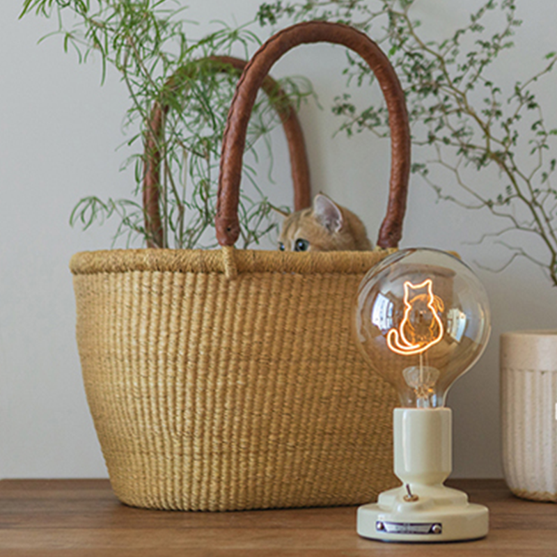 Pale-base cat-outline lamp on a wood console beside a woven basket with a cat and greenery, near a pale ribbed planter