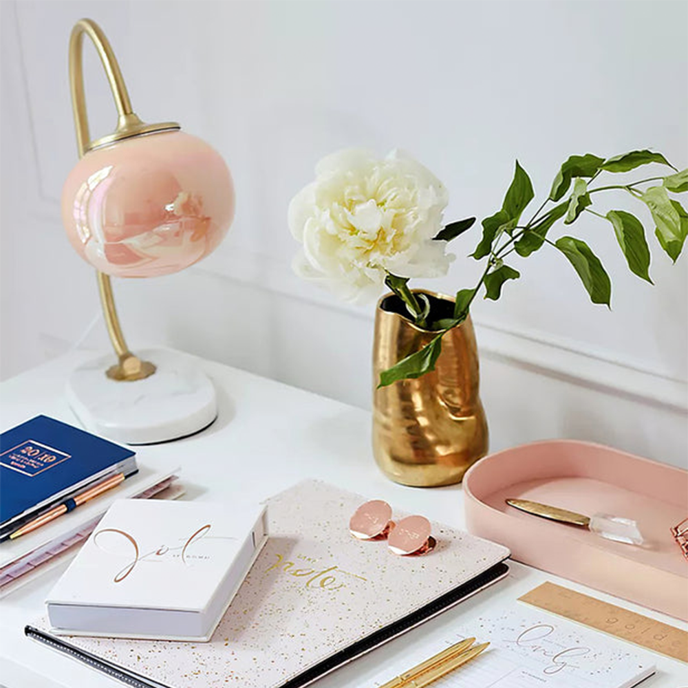 Pink glass globe lamp on a white desk beside a gold-tone flower vase, notebooks, pens and a pink tray