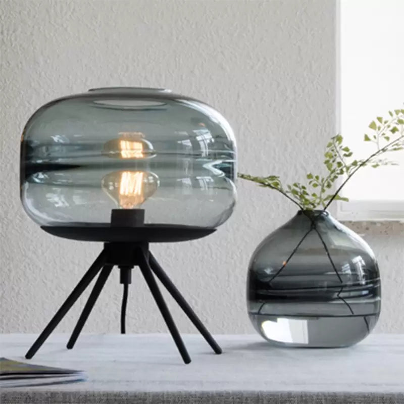 Blue-tinted glass lamp on black splayed legs beside a rounded vase with green branches near a window