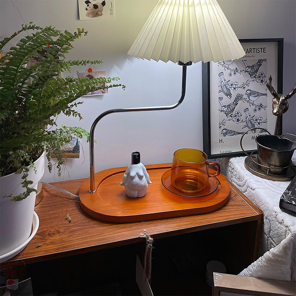 Lit pleated lamp on a wooden cabinet beside a fern, amber cup, white figurine bottle and framed bird print