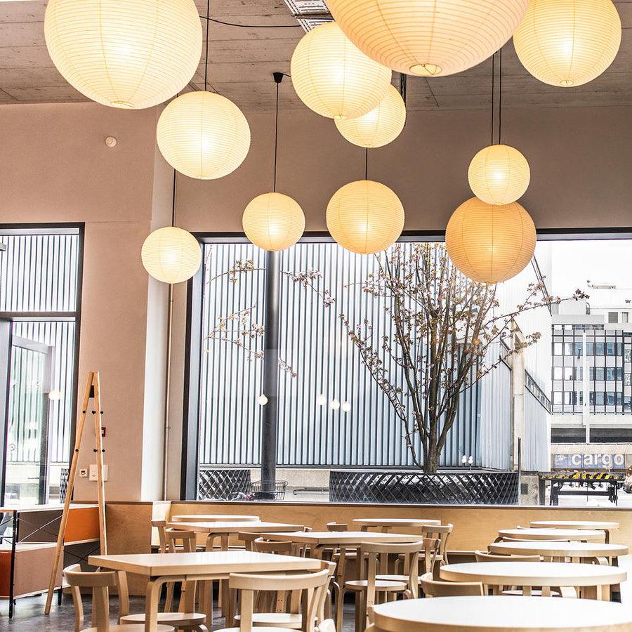 Cafe side view of glowing paper globes above wood tables beside wide windows and a flowering tree