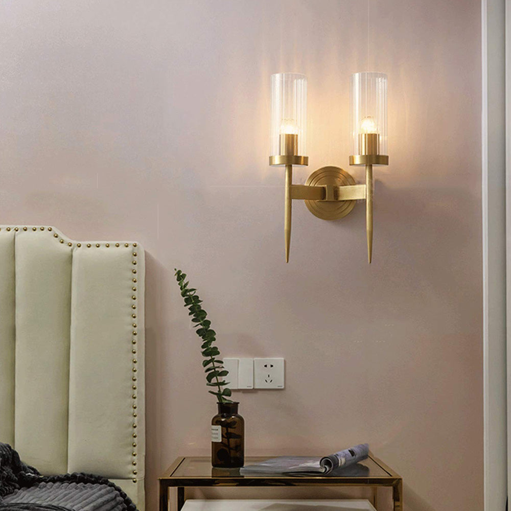 Double-arm gold-tone wall light with two ribbed sleeves beside a headboard and table with a vase and rolled pages