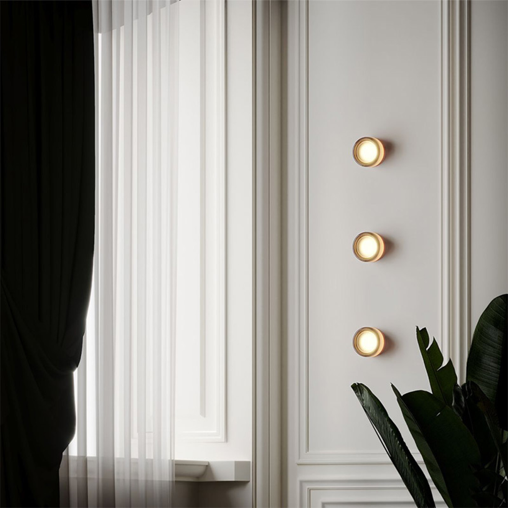 Three amber-looking round wall lights in a vertical row beside sheer curtains and green leaves