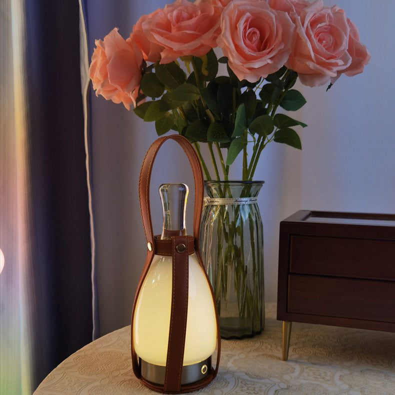 Brown leather-handle Bell lamp on a cream patterned table beside a glass vase of pink roses and a small wooden drawer unit