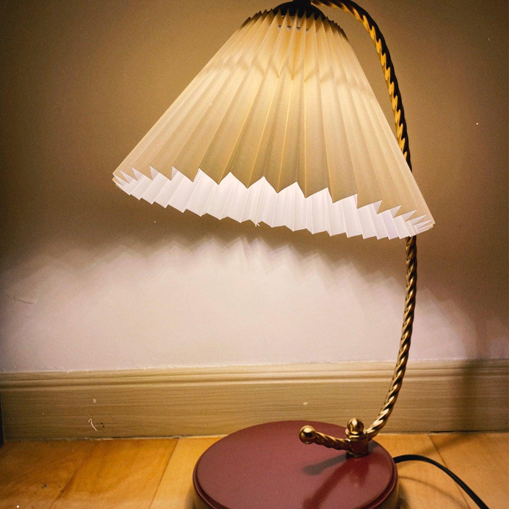 Lit Beige-looking pleated shade and gold-tone twisted stem above a brown-red base near wood trim