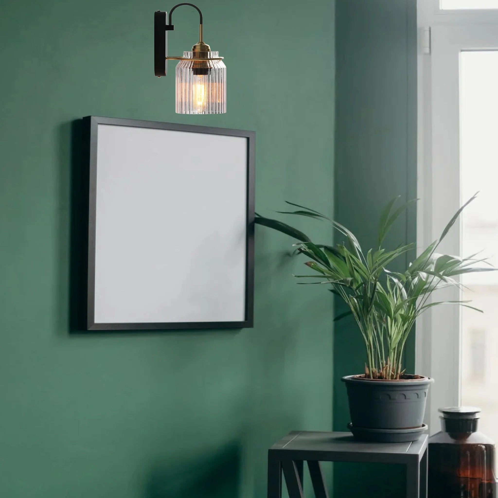 Clear ribbed glass wall light above a black picture frame on a green wall beside a window and potted palm