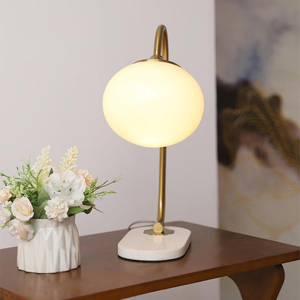 Front view of a lit pale globe lamp and oval base on a wood table beside a white vase and framed art